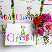 The Cheerful Baker packaging with colorful branding and a vase of flowers on a white background