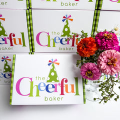 The Cheerful Baker packaging with colorful branding and a vase of flowers on a white background